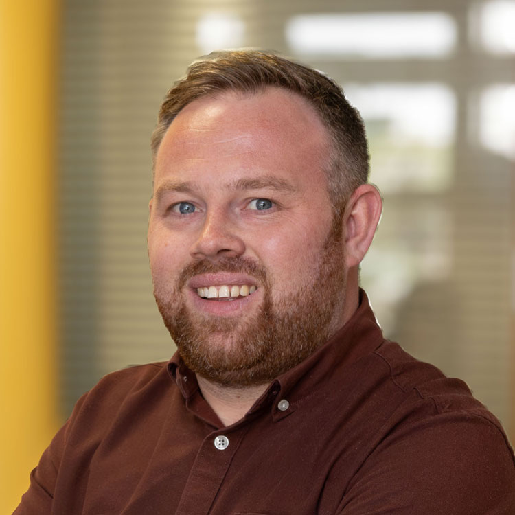 A man sporting a beard and a brown shirt, facing the viewer with a relaxed posture and expression.