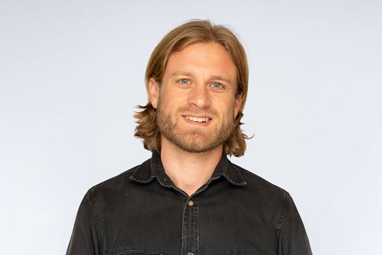A man with long hair dressed in a black shirt, posing confidently.