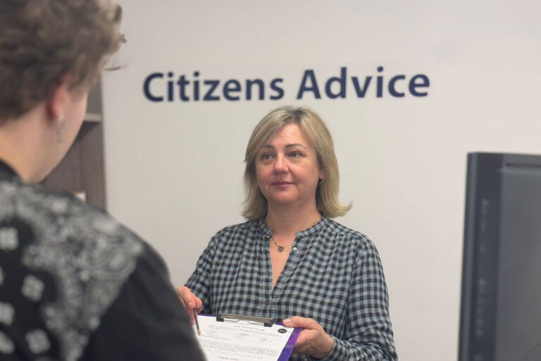 A woman with blonde hair, wearing a chequered blouse, smiles while holding a clipboard in an office setting.