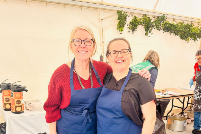 Two women in aprons stand side by side, smiling and engaged in conversation in a kitchen setting.
